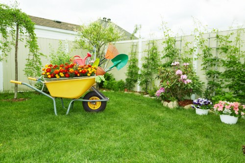 A well-maintained backyard garden featuring a lush, evenly mowed green lawn bordered by flower beds with blooming pink, purple, and white flowers in white pots. A yellow wheelbarrow filled with colorful garden flowers and tools such as a watering can and small spade are positioned on the grass, indicating ongoing garden care activities. The garden is enclosed by a white wooden fence covered in green climbing plants and small shrubs, creating a private and inviting outdoor space within a residential area. In the background, a small tree with light green foliage and a neatly trimmed hedge line the fence, adding natural structure and greenery to the scene under a slightly overcast sky. The environment appears bright and fresh, suitable for professional gardening and lawn maintenance services offered by Lawn Mowing Golders Green in the London NW11 area, reflecting a typical suburban garden in Golders Green.
