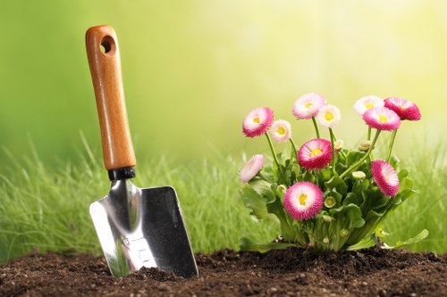 A close-up view of a garden with freshly turned dark soil in the foreground, where a small gardening trowel with a wooden handle is embedded upright, partially covered by soil. To the right, there is a vibrant cluster of pink and white flowering plants with rounded blooms and green leaves, suggesting a decorative flower bed within a landscaped garden area. The background features a lush, green lawn with soft natural light illuminating the scene, emphasizing the healthy grass and the seasonal outdoor environment typical of gardens in Golders Green, London. The setting appears well-maintained and suitable for professional gardening or lawn care services offered by Lawn Mowing Golders Green, with a focus on outdoor landscaping, planting, and turf management in residential gardens.